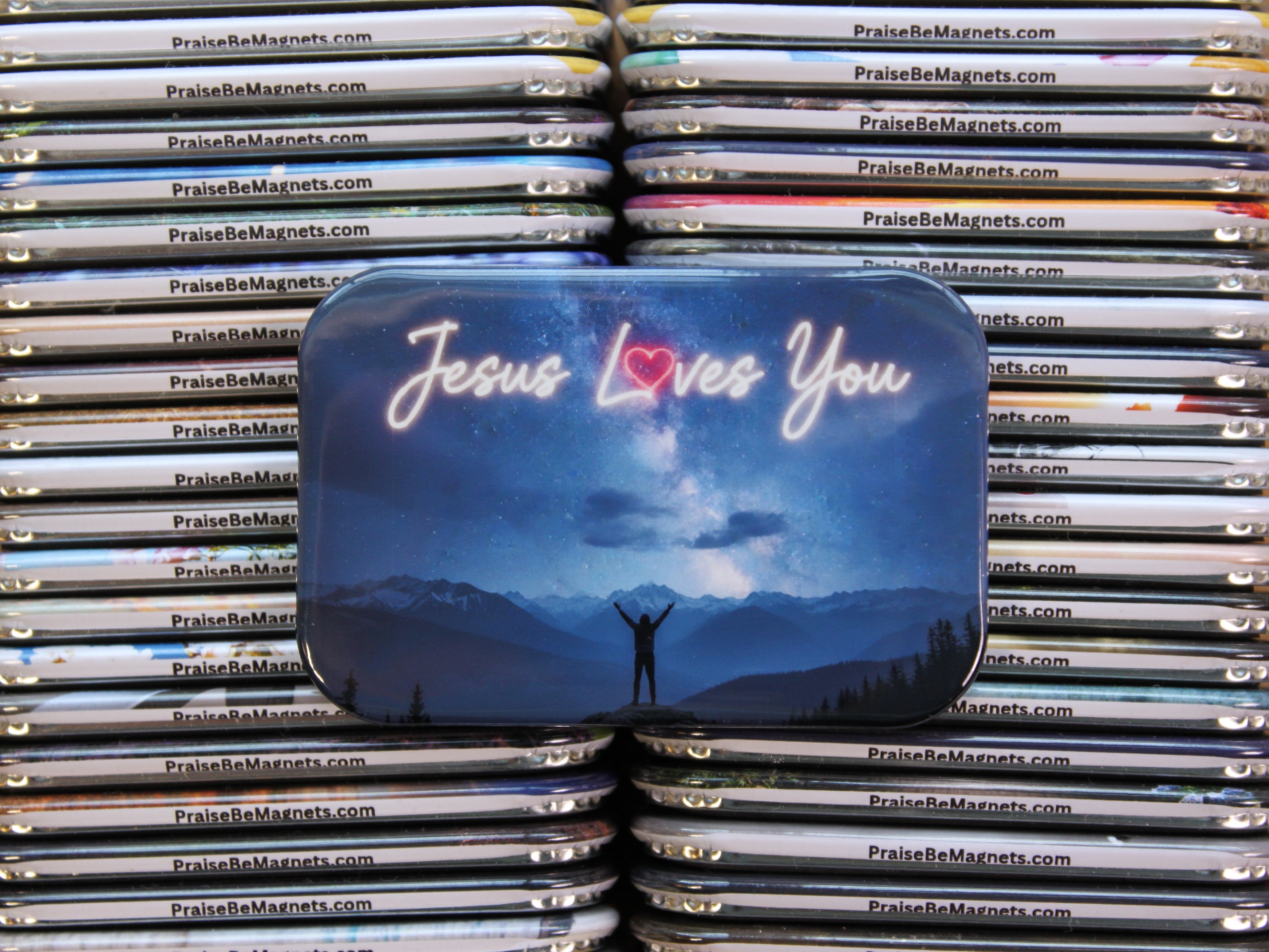 Stack of 'Praise Be Magnets' with a prominent 'Jesus Loves You' tin in the foreground.