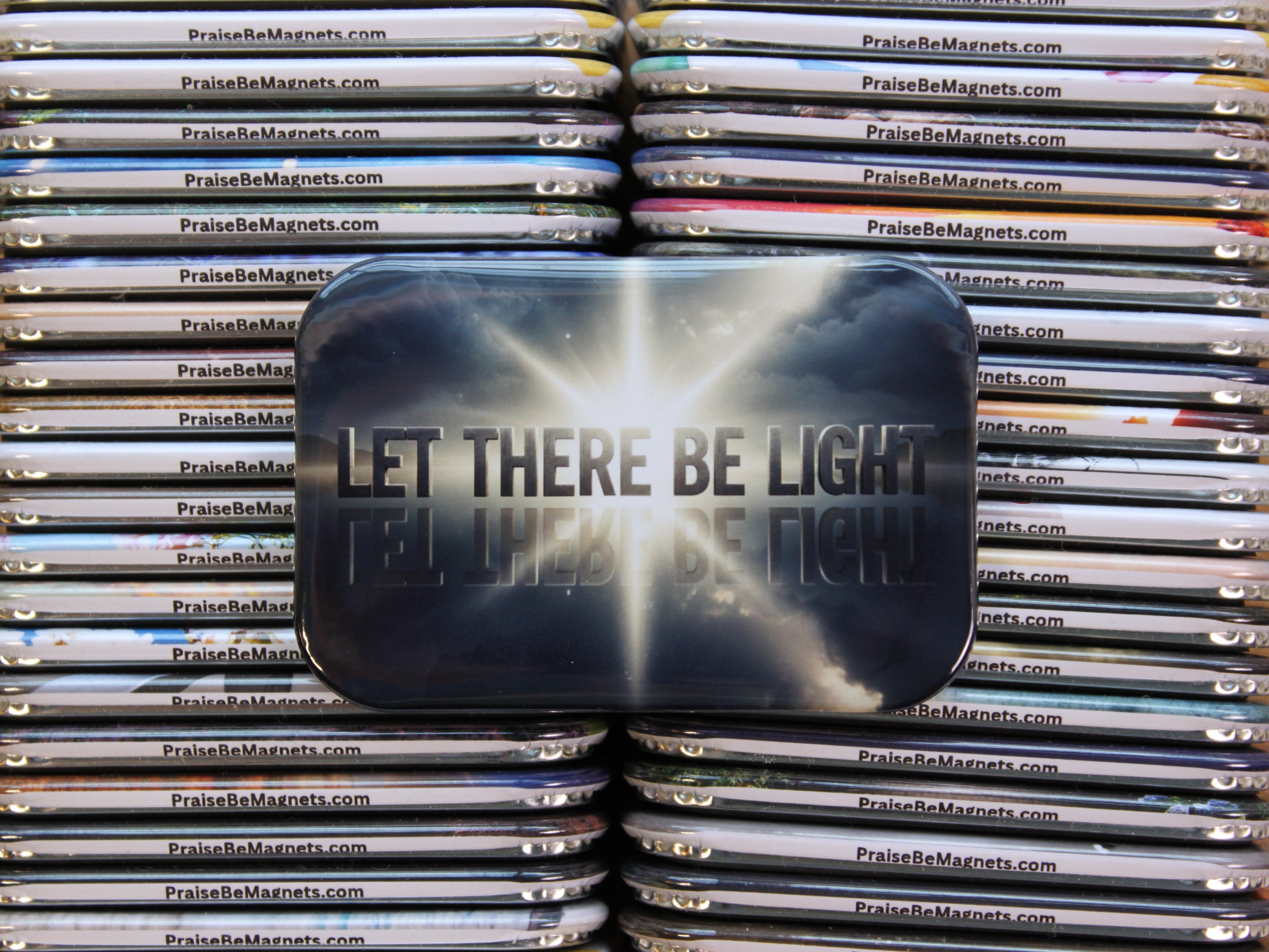 Stack of metal magnets with 'PraiseBeMagnets.com' branding, featuring a prominent magnet with 'LET THERE BE LIGHT' text.