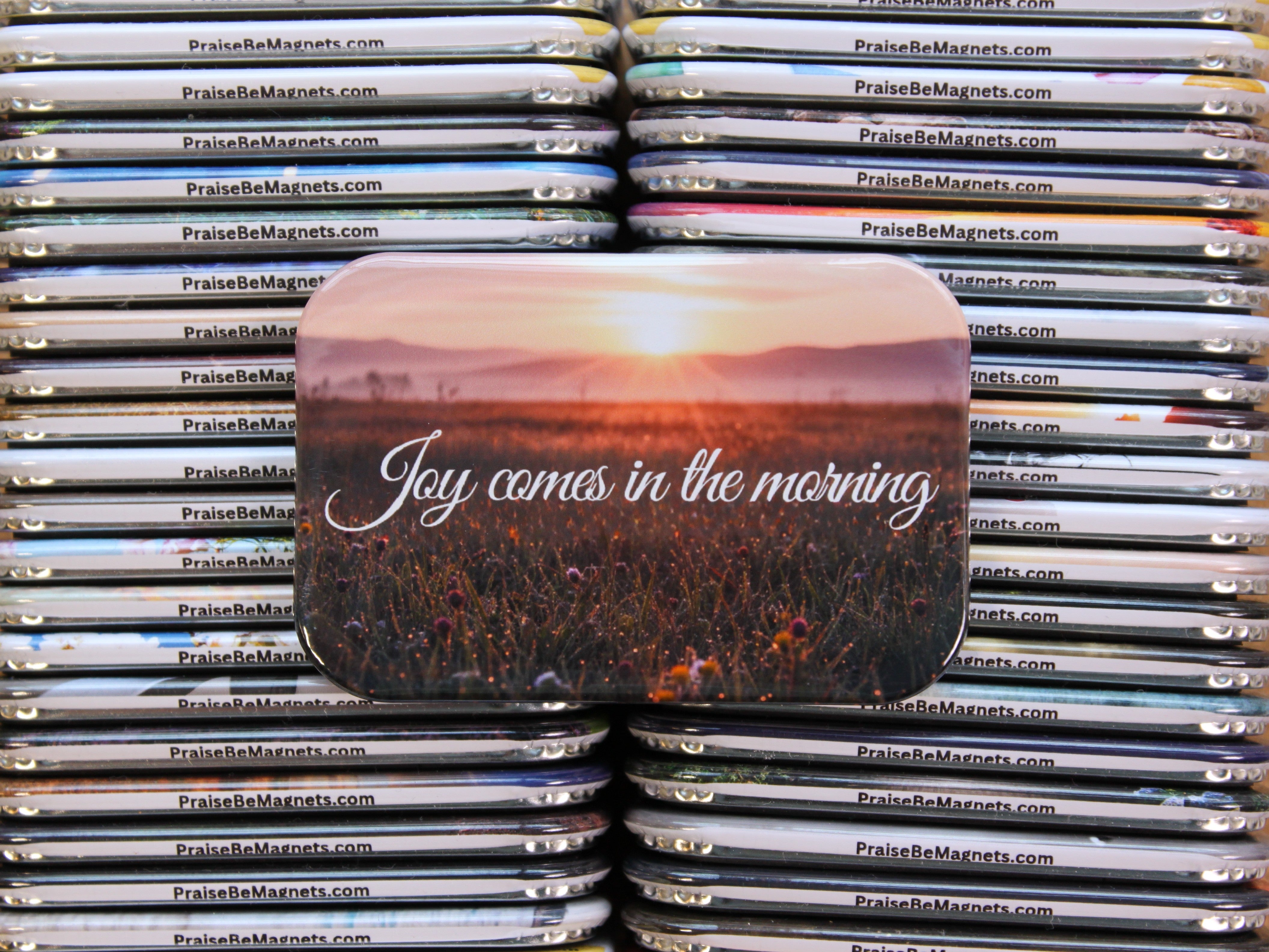 Stack of 'PraiseBeMagnets' products with a central magnet featuring 'Joy comes in the morning' text.