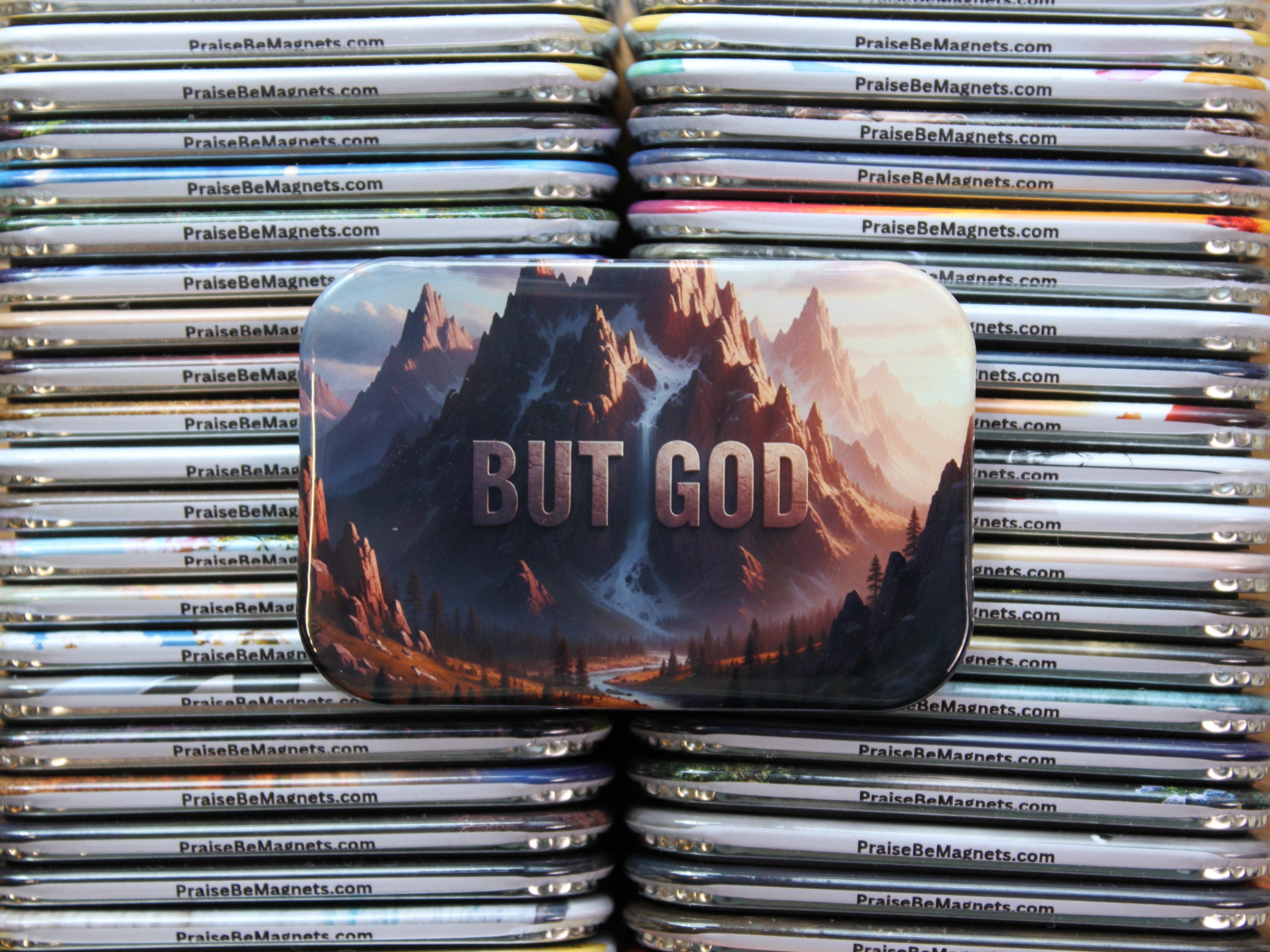 Stack of metal magnets with mountain design and 'But God' text, branded 'PraiseBeMagnets'.