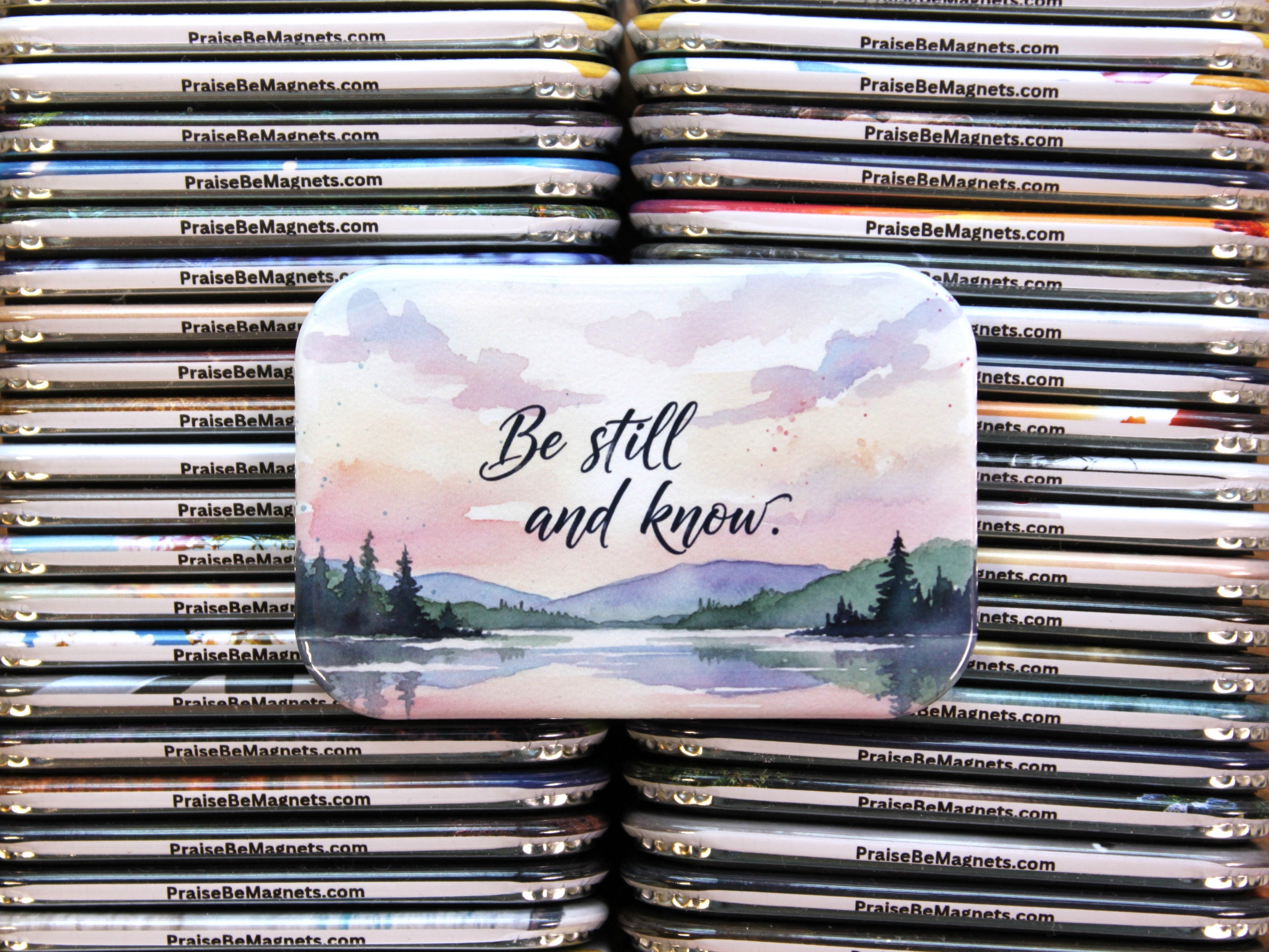 Stack of 'Praise Be Magnets' with a scenic magnet featuring 'Be still and know.' text.
