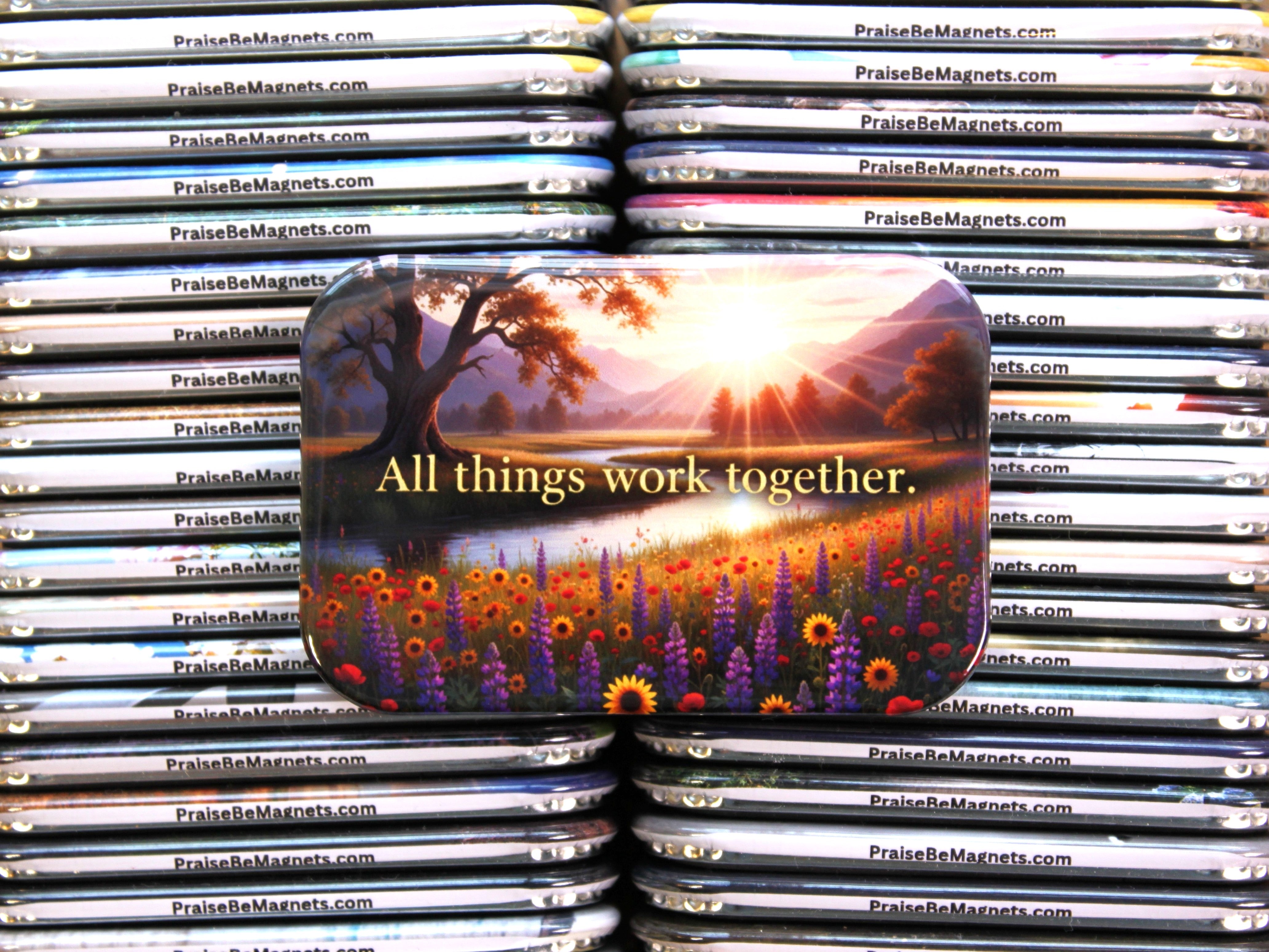 Stack of PrayBeMagnets with a prominent magnet featuring a sunset and 'All things work together' text.