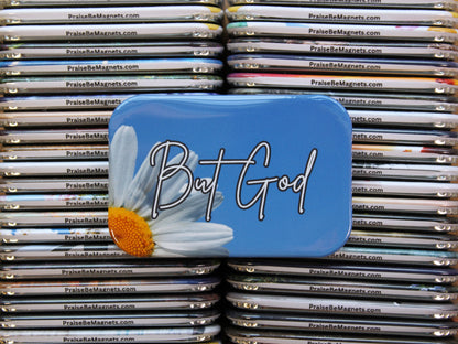 Blue magnet with 'But God' text and daisy design on a stack of similar magnets.